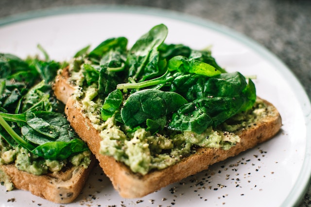 Two slices of brown bread topped with mashed avocados and spinach leaves, served on a ceramic plate. 