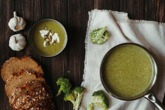 A pot of green soup with a bowl filled with the same, sliced bread, broccoli bits, and garlic on the side.