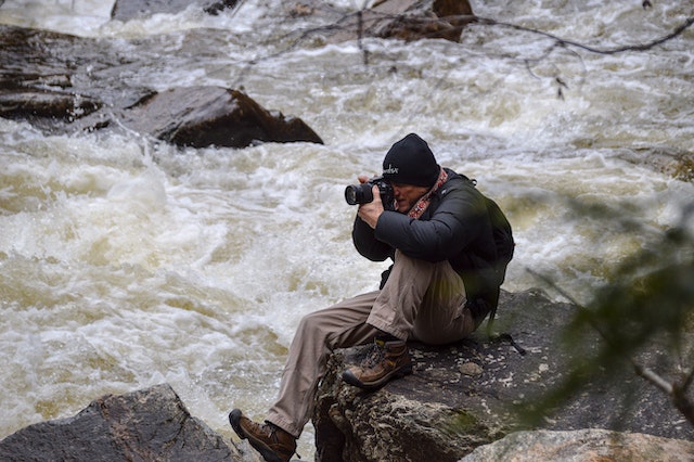  A man sitting on a boulder beside a raging river while shooting a camera