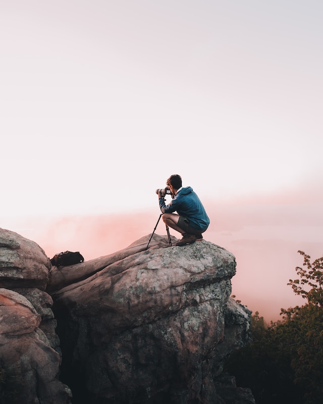 Man in blue jacket with a camera and tripod set up on top of a rocky cliff