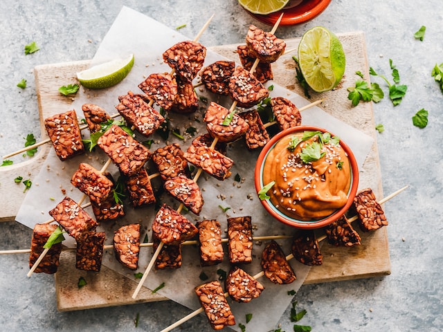 Skewered tempeh served on a wooden board with a side of orange-looking dip
