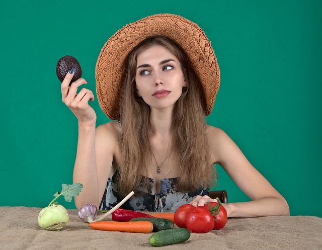 Woman in a hat holding an avocado with her hand, several fruits and vegetables on the table before here